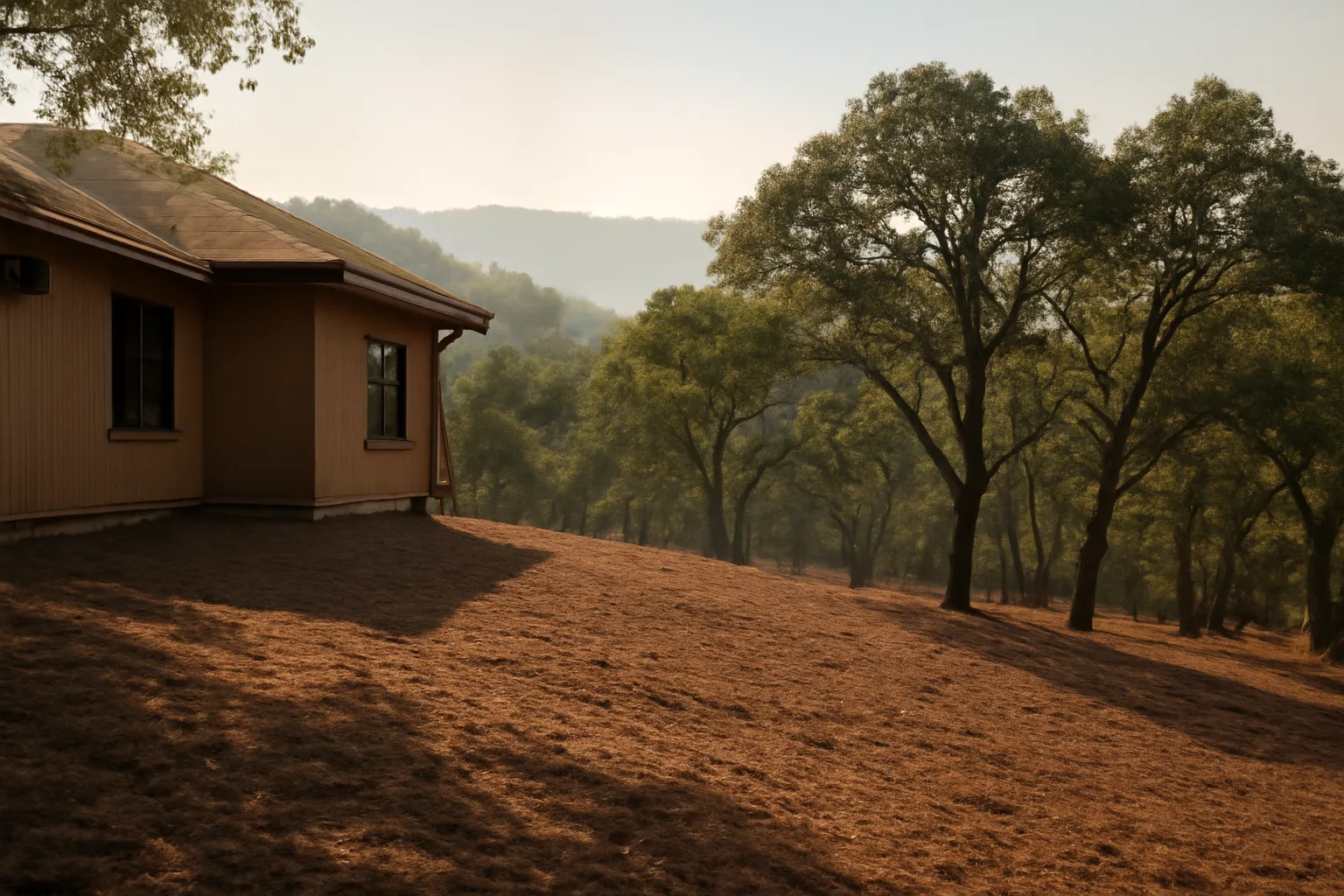 Cleared defensible space zone around a hillside home with thinned oak trees and distant smoky ridge